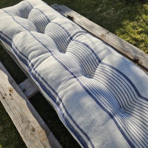Può includere: Un cuscino a righe bianche e blu con bottoni. Il cuscino è realizzato in tessuto di lino ed è perfetto per aggiungere un tocco di stile a qualsiasi panca o area salotto all'aperto.