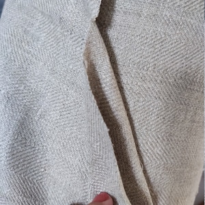 May include: Close-up of a beige herringbone fabric with a textured weave. The fabric is folded over, showing the underside.