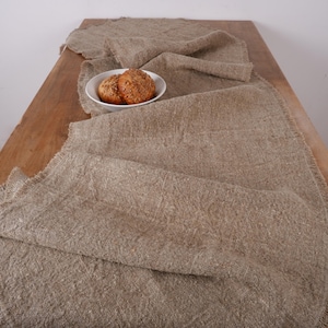 Puede incluir: Un camino de mesa r&uacute;stico de lino beige con borde deshilachado, sobre una mesa de madera. Un cuenco blanco contiene dos panecillos redondos con semillas. La textura natural del lino y la madera crea una escena c&aacute;lida y acogedora.