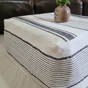 May include: A rectangular ottoman with a cream-colored fabric and black horizontal stripes. A small brown vase with a floral design holds a bouquet of blue and white flowers. The ottoman is placed in front of a brown leather sofa.