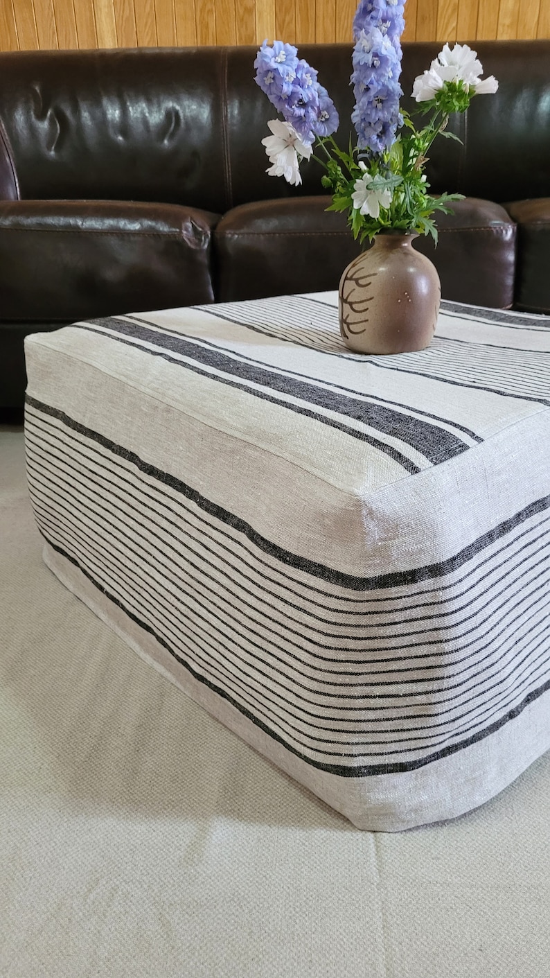 May include: A rectangular ottoman with a cream-colored fabric and black horizontal stripes. A small brown vase with a floral design holds a bouquet of blue and white flowers. The ottoman is placed in front of a brown leather sofa.