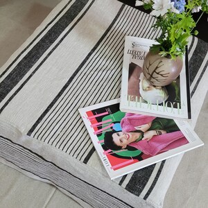 May include: A rectangular cushion with a cream-colored background and black stripes. Two fashion magazines and a small vase with flowers rest on top. The magazines have colorful covers. The vase is brown and has a leaf design.