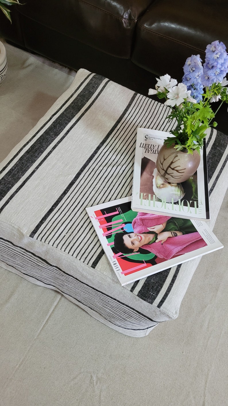 May include: A rectangular cushion with a cream-colored background and black stripes. Two fashion magazines and a small vase with flowers rest on top. The magazines have colorful covers. The vase is brown and has a leaf design.