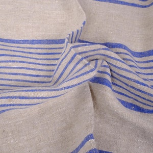 May include: A close-up of a beige linen fabric with blue stripes. The fabric is folded and wrinkled, showing the texture of the material.