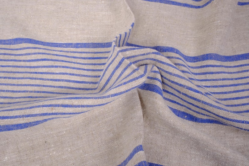 May include: A close-up of a beige linen fabric with blue stripes. The fabric is folded and wrinkled, showing the texture of the material.