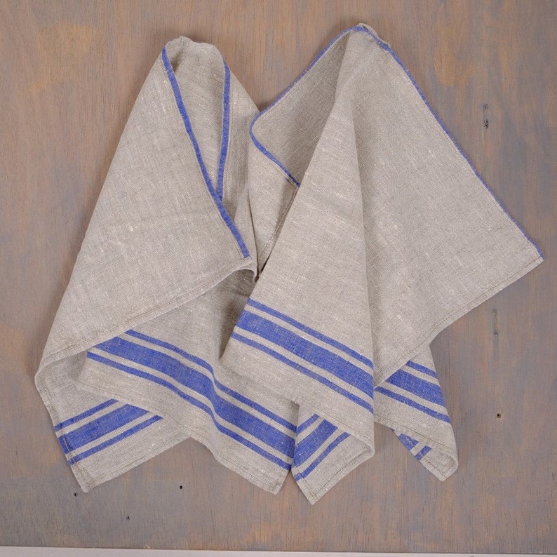 May include: Two linen tea towels with blue stripes. The towels are folded and draped over a wooden surface.