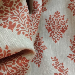 May include: Close-up of a beige linen fabric with a repeating pattern of rust-colored floral motifs. The fabric is folded over, showing the pattern on both sides.