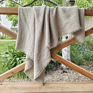 Peut inclure: Une couverture en lin beige avec une bordure effilochée est drapée sur une clôture en bois. La couverture a un aspect texturé et est suspendue en plis souples. L'arrière-plan présente de la verdure et un arbre, suggérant un cadre extérieur.