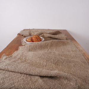 Puede incluir: Un camino de mesa de lino beige con borde deshilachado, sobre una mesa de madera. Un cuenco blanco contiene dos productos horneados redondos de color marr&oacute;n dorado. La escena evoca calidez y simplicidad natural, ideal para un comedor acogedor.