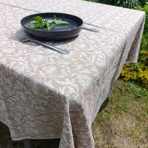 May include: A rectangular tablecloth featuring a light beige and white botanical pattern of leaves and flowers. A dark grey ribbed bowl containing green herbs and a silver metal fork are placed on the surface of the fabric.