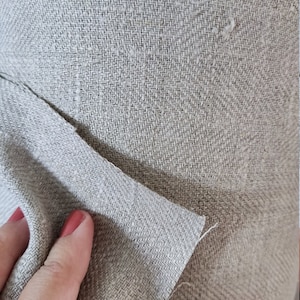 May include: Close-up of a beige linen fabric with a woven texture. The fabric is folded over itself, showing the underside of the material.