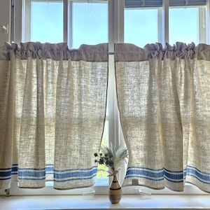 Op de afbeelding: Linnen café gordijnen in een natuurlijke beige tint met blauwe strepen aan de onderkant. De gordijnen hangen voor een raam, met een kleine vaas met bloemen op de vensterbank. De gordijnen hebben een geplooid bovenstuk.