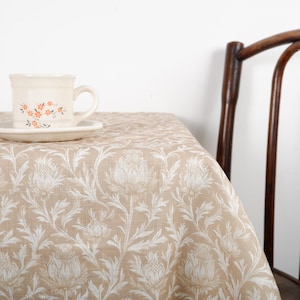 May include: A beige tablecloth with a white floral design, with a teacup and saucer featuring orange floral details. A wooden chair is partially visible. The tablecloth is covering a table.