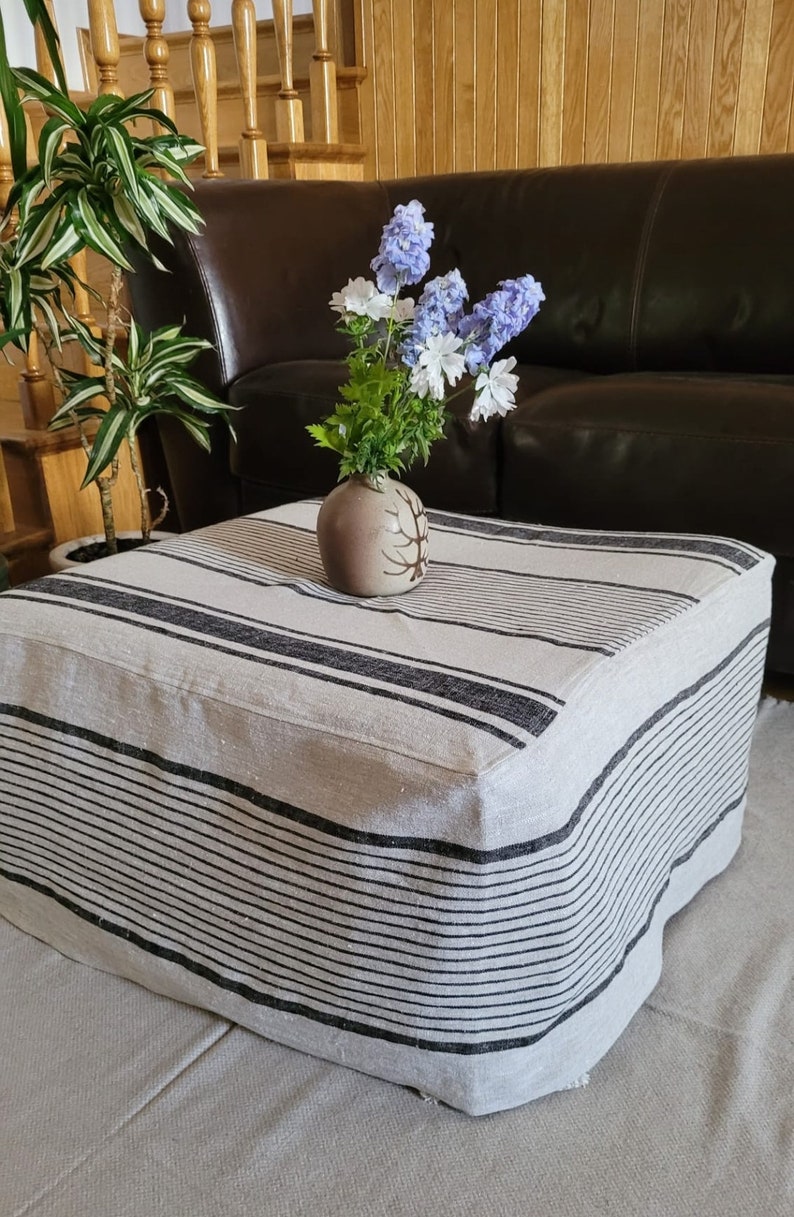 May include: A square ottoman with a striped fabric cover in black and off-white. A small, round, tan vase with a tree design holds blue and white flowers. The ottoman is in front of a brown leather sofa.