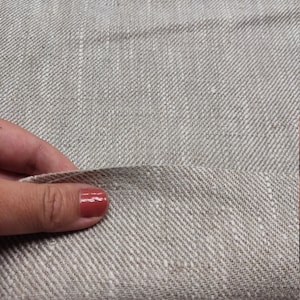May include: Close-up of a beige linen fabric with a subtle herringbone weave. The fabric is folded in half and a hand is holding the fold.