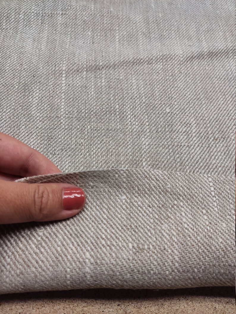 May include: Close-up of a beige linen fabric with a subtle herringbone weave. The fabric is folded in half and a hand is holding the fold.
