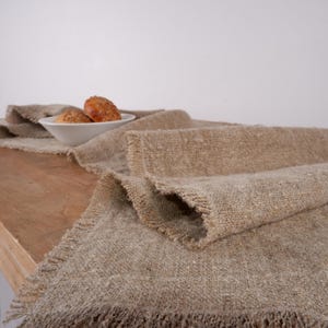 Puede incluir: Un camino de mesa de lino beige con borde deshilachado, sobre una superficie de madera. Un cuenco blanco contiene dos panecillos dorados. La textura y el color del camino de mesa crean una est&eacute;tica r&uacute;stica y acogedora.