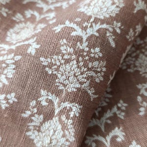 Pode incluir: Close-up de um tecido castanho com um padrão floral branco. O tecido tem uma superfície texturizada e um brilho sutil.