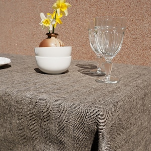 Op de afbeelding: Een eettafel met een bruin en wit visgraatpatroon tafelkleed. Twee heldere waterglazen, twee witte kommen en een kleine vaas met gele en witte narcissen zijn op de tafel geplaatst.