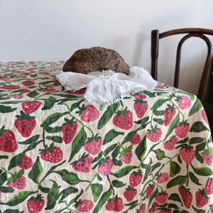 Puede incluir: Un mantel blanco con estampado de fresas. El mantel está sobre una mesa con una hogaza de pan encima.