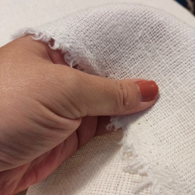 Puede incluir: Primer plano de una tela blanca con un borde deshilachado. La tela es texturizada y tiene un aspecto tejido. Una mano sostiene la tela, y una u&ntilde;a est&aacute; pintada de color naranja claro.