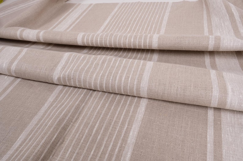 May include: Close-up of a beige linen fabric with white stripes. The fabric is folded and has a textured surface.