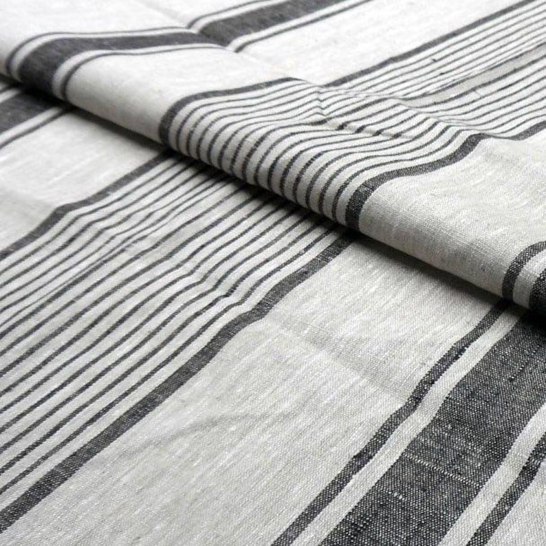 May include: A close-up of a white linen fabric with black stripes. The fabric is folded in half, showing the texture of the linen.