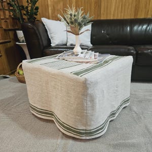 Könnte beinhalten: Ein quadratischer Ottoman mit einem Bezug aus Leinen mit grünen und weißen Streifen. Der Ottoman steht auf einem grauen Teppich vor einem braunen Ledersofa.
