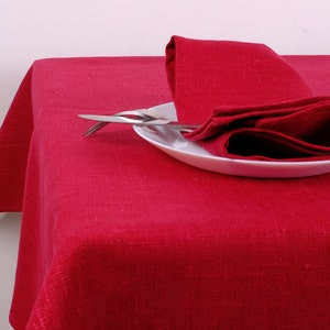 Op de afbeelding: Een rode linnen tafelkleed bedekt een tafel, met een bijpassende gevouwen servet en bestek op een wit bord. Het tafelkleed is effen, levendig rood van kleur en suggereert een eethema.