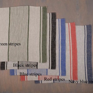 May include: A collection of linen napkins with vertical stripes in various colors: green, black, blue, red, and navy blue. The napkins are arranged in a layered display, showcasing the different stripe patterns and colors.