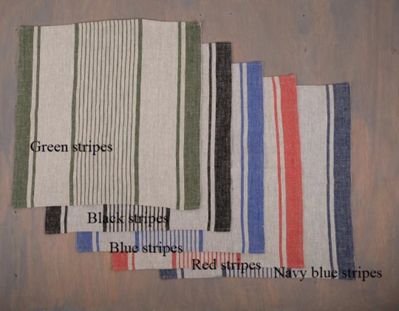 May include: A collection of linen napkins with vertical stripes in various colors: green, black, blue, red, and navy blue. The napkins are arranged in a layered display, showcasing the different stripe patterns and colors.