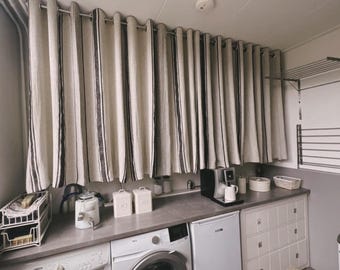 May include: A white kitchen with a washing machine, a dryer, and a set of white cabinets. The kitchen has a window with striped curtains and a countertop with a coffee maker and other appliances.