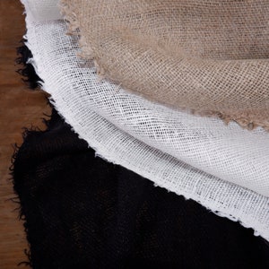 May include: Three layers of fabric with raw, frayed edges stacked on top of each other. The top layer is a natural tan burlap, the middle layer is a white open-weave mesh fabric, and the bottom layer is a solid black woven textile material.