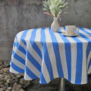 May include: A round tablecloth with alternating blue and white stripes covers a table. A white vase with greenery and a teacup with a floral design sit on top. The tablecloth is made of a soft fabric.