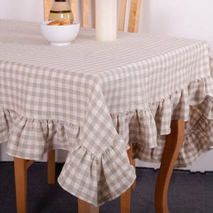 Può includere: Una tovaglia a quadri beige e bianca con bordo a balze. La tovaglia è drappeggiata su un tavolo di legno, con una ciotola bianca, una candela e una bottiglia decorativa visibili sopra. Il tema generale è l'arredamento della casa.