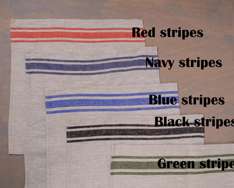 May include: Five linen tea towels with different colored stripes. The towels are arranged in a stack, with the red striped towel on top and the green striped towel on the bottom. The towels are labeled with the color of their stripes: red, navy, blue, black, and green.