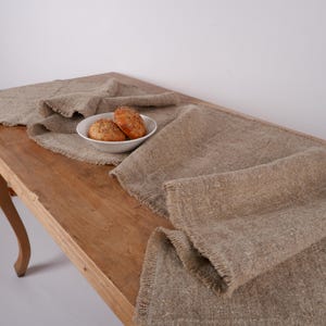 Puede incluir: Un camino de mesa r&uacute;stico de lino en color beige natural con borde deshilachado. Dos panecillos redondos est&aacute;n en un cuenco blanco sobre una mesa de madera. El camino de mesa est&aacute; drapeado sobre la mesa.