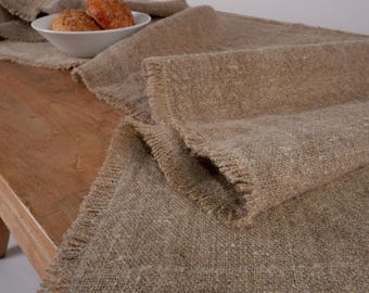 May include: A rustic, woven table runner in shades of tan and beige, with a fringed edge. The runner is draped over a wooden table, with a white bowl holding two rolls in the background. The texture of the fabric is visible.