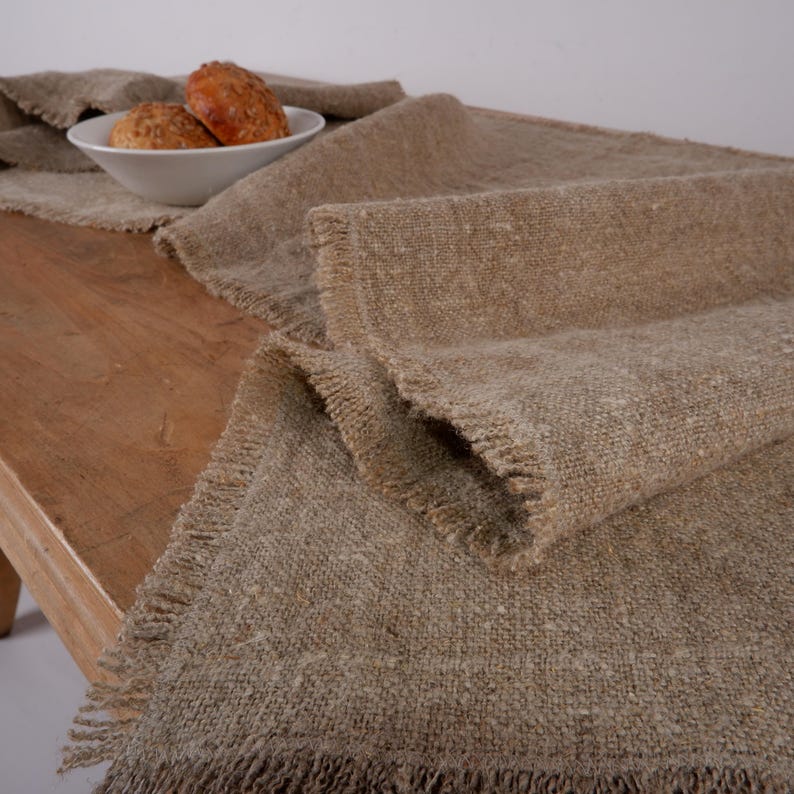 Puede incluir: Un camino de mesa r&uacute;stico, tejido en tonos beige y marr&oacute;n claro, con bordes deshilachados. Est&aacute; colocado sobre una mesa de madera, con un cuenco blanco que contiene dos panecillos en el fondo. La textura del tejido es visible.