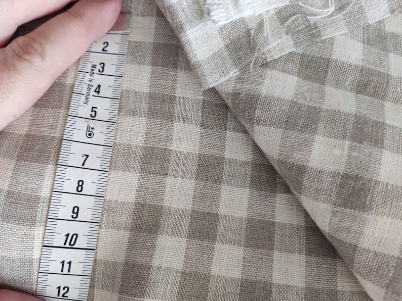 May include: A close-up of a beige and white gingham fabric with a measuring tape showing 12 inches. The fabric is folded over itself, revealing the pattern.