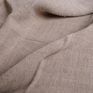 May include: Close-up of a natural beige linen fabric with a textured weave. The fabric is folded and draped, creating soft folds and wrinkles.