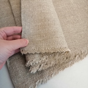 Puede incluir: Una pila de tela de lino natural con bordes deshilachados. La tela es de color beige claro y tiene una textura tejida.