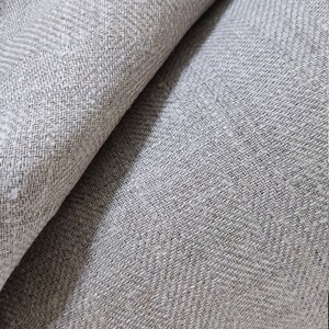 May include: Close-up of a light gray fabric with a subtle herringbone pattern. The fabric is woven with a textured, slightly rough surface.