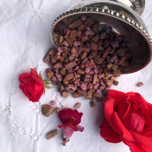 Op de afbeelding: Een zilveren kom die overloopt met kleine, ruwe, roze en bruine edelstenen. De kom is omgevallen en de edelstenen vallen op een witte stoffen achtergrond. Twee rode rozen en een roze bloem zijn rond de kom verspreid.