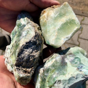 May include: Three rough-cut, raw green stones with a translucent quality. The stones have a mix of light and dark green hues, with some areas appearing almost white. The texture is rough and uneven, with visible mineral formations.