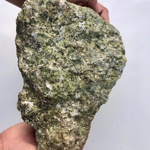 Pode incluir: Uma rocha grande, de forma irregular, com uma superfície áspera e texturizada. A rocha exibe uma mistura de cores verdes, brancas e cinza escuro, criando uma aparência manchada. A rocha é segurada em duas mãos contra um fundo branco.