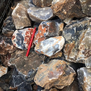 Puede incluir: Una pila de piedras de ágata en bruto, grises, marrones y blancas con bandas rojas y blancas. Las piedras son de varios tamaños y formas.