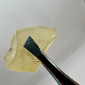 Może przedstawiać: Mały, nieregularny, przezroczysty żółto-zielony obiekt jest trzymany przez metalowe narzędzie. Obiekt ma lekko szorstką teksturę i wydaje się być fragmentem czegoś większego. Narzędzie jest srebrne i ma płaską, fazowaną krawędź.