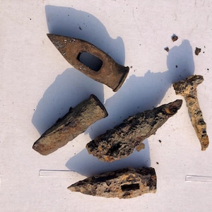 May include: Five rusty metal objects, including a hammer, a nail, and three chisels, are arranged on a white surface. The objects are covered in rust and dirt.