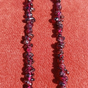 May include: Two strands of dark red gemstone beads, possibly garnet, strung on a clear thread. The beads are irregular in shape and size, and they are arranged in a loose, flowing pattern.
